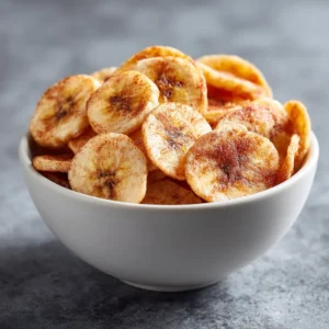 A minimalist white bowl filled with homemade banana chips, showcasing their golden-brown color and crispy texture against a clean background.