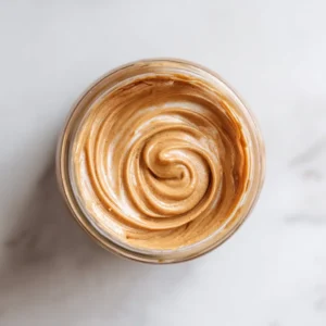 An overhead shot of homemade cookie butter in a clear glass jar, with a spoon resting on the side and Biscoff cookies scattered around.