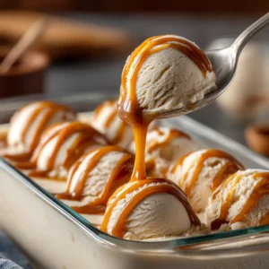 Close up of a metal spoon lifting a scoop of creamy beige salted caramel ice cream from a clear glass loaf pan.