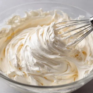 Close-up of thick, snow-white Swiss Meringue Buttercream inside a clear glass mixing bowl with a balloon whisk.