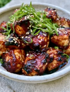 Close-up shot of bite-sized chicken breast cubes coated in thick, glossy dark BBQ sauce with charred edges and sesame seed garnish. (Honey BBQ Chicken Bites)
